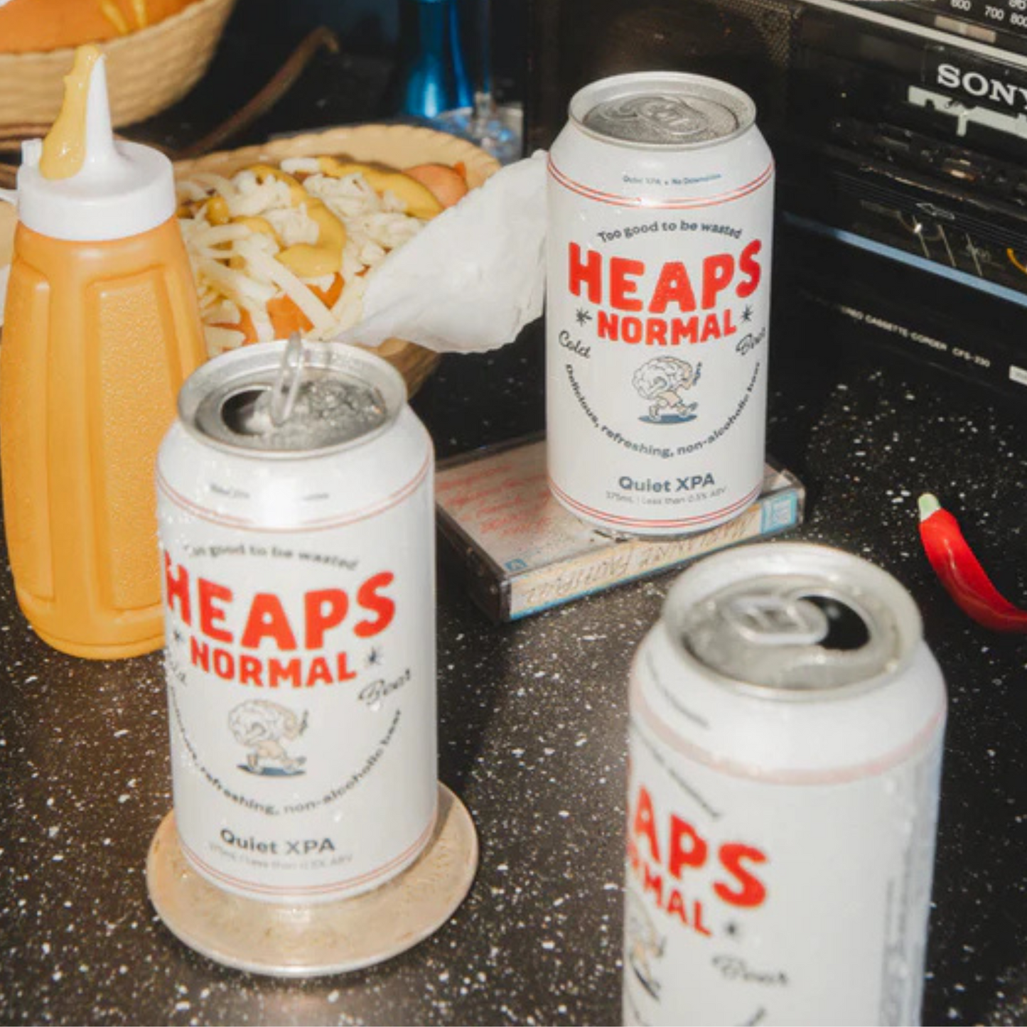 Three cans of 'Heaps Normal' Quiet XPA on a table with condiments and a radio in the background.