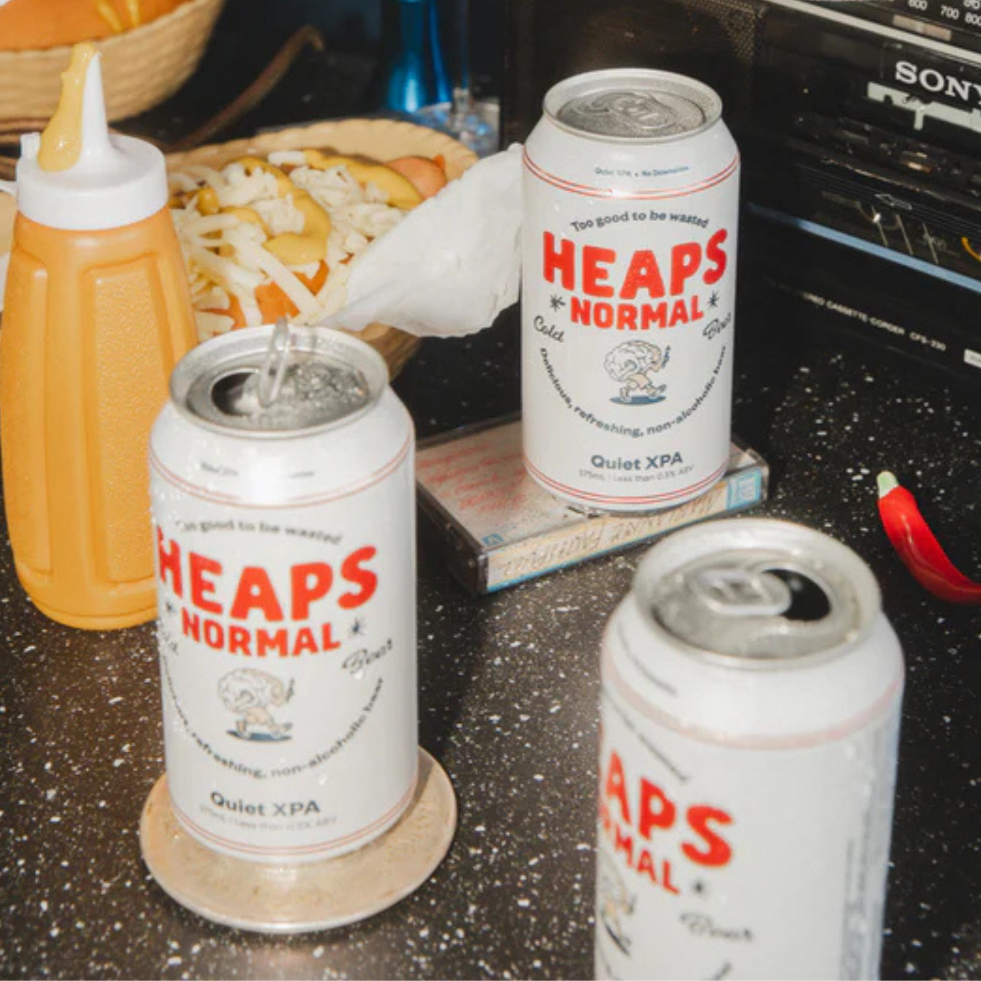 Three cans of 'Heaps Normal' Quiet XPA on a table with condiments and a radio in the background.