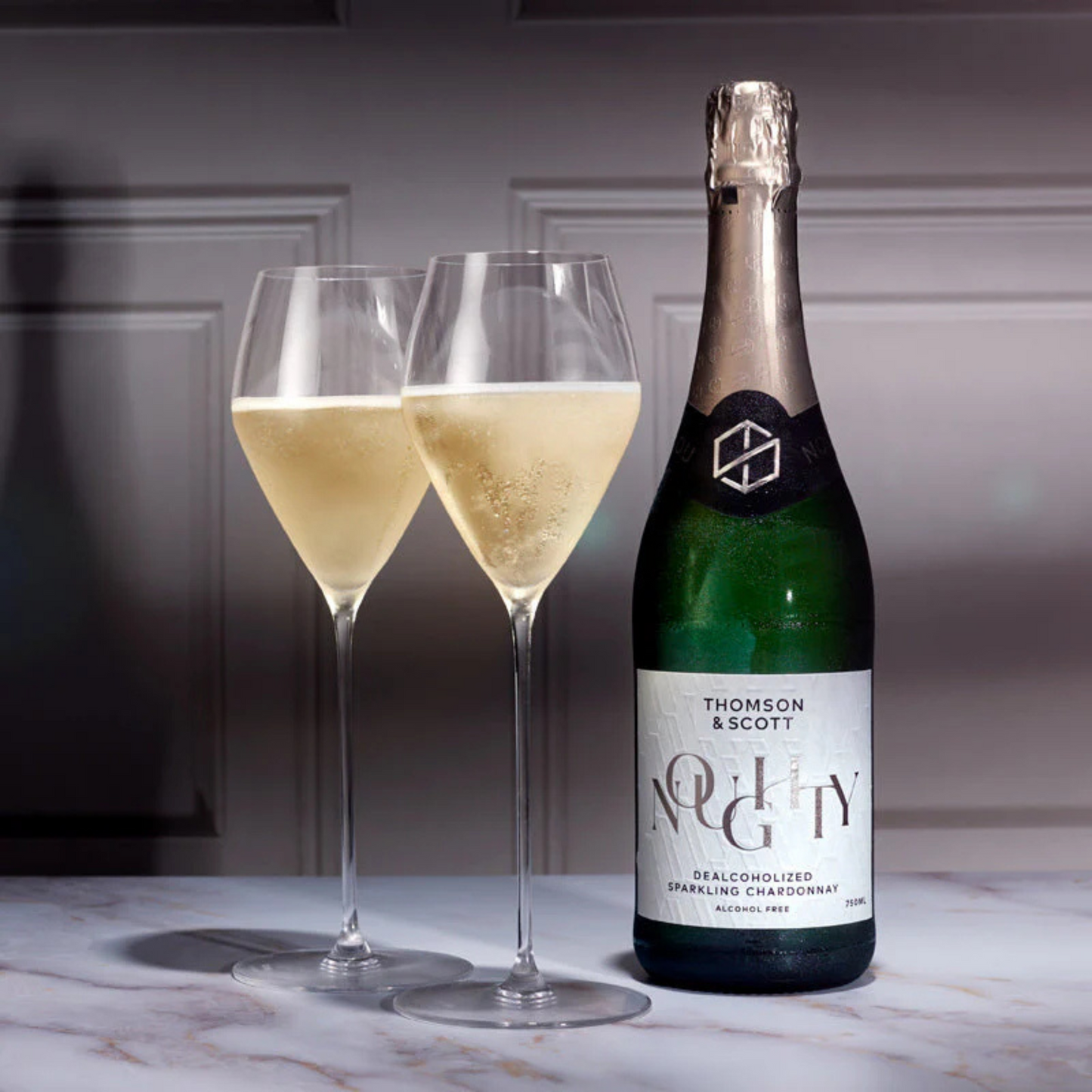 Bottle of Thomson & Scott Noughty sparkling wine with two filled glasses on a marble surface.