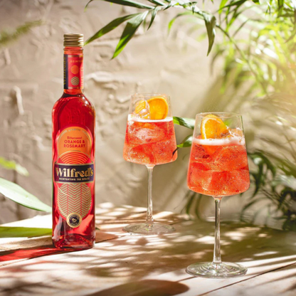 Bottle of Wilfred's pink cocktail with two glasses filled with the same cocktail on a wooden surface with greenery in the background.