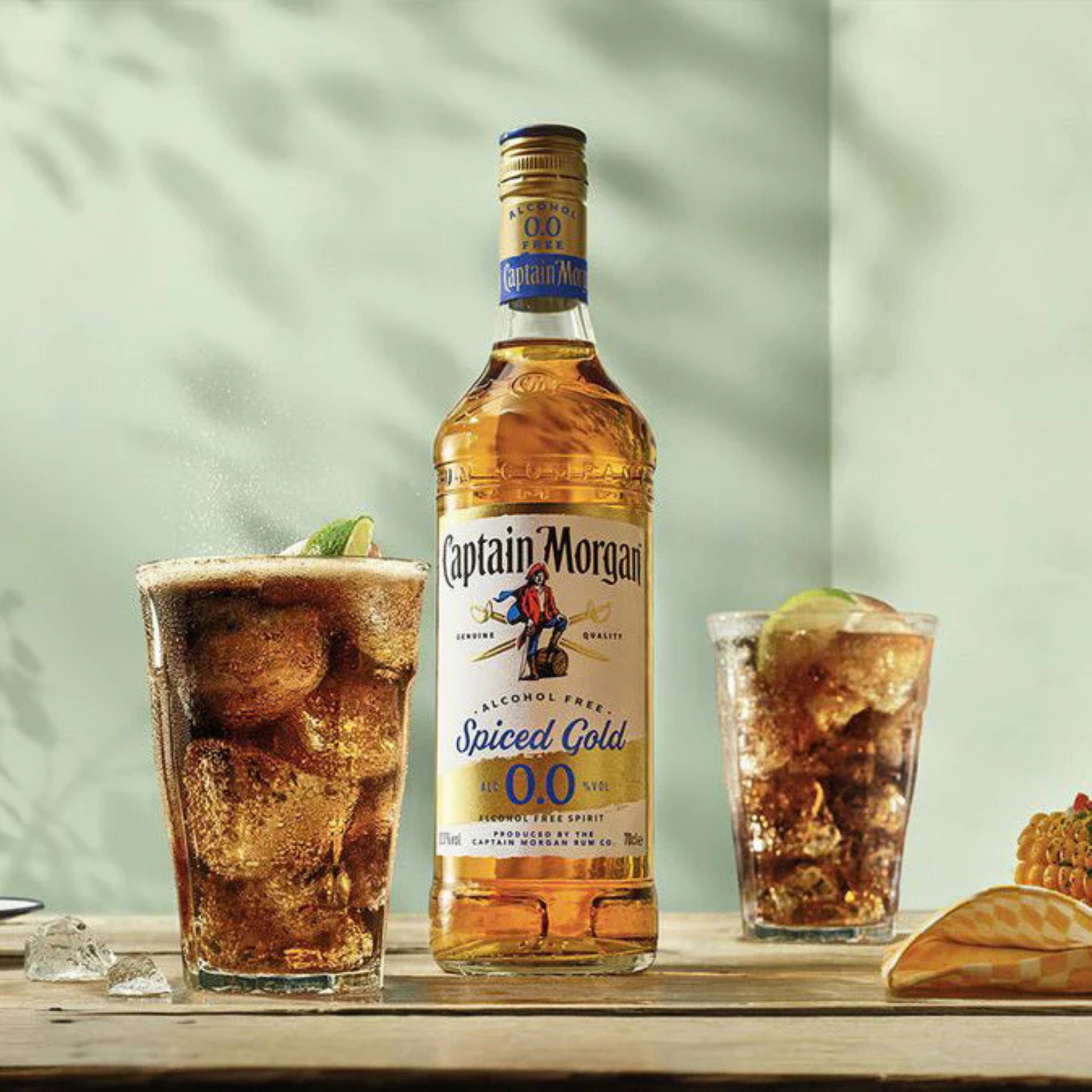 Captain Morgan Spiced Gold bottle with two glasses of a mixed drink on a wooden surface.