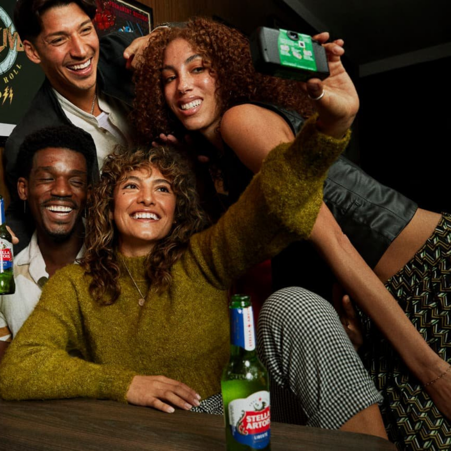 Group of friends taking a selfie in a bar with Stella Artois bottles.
