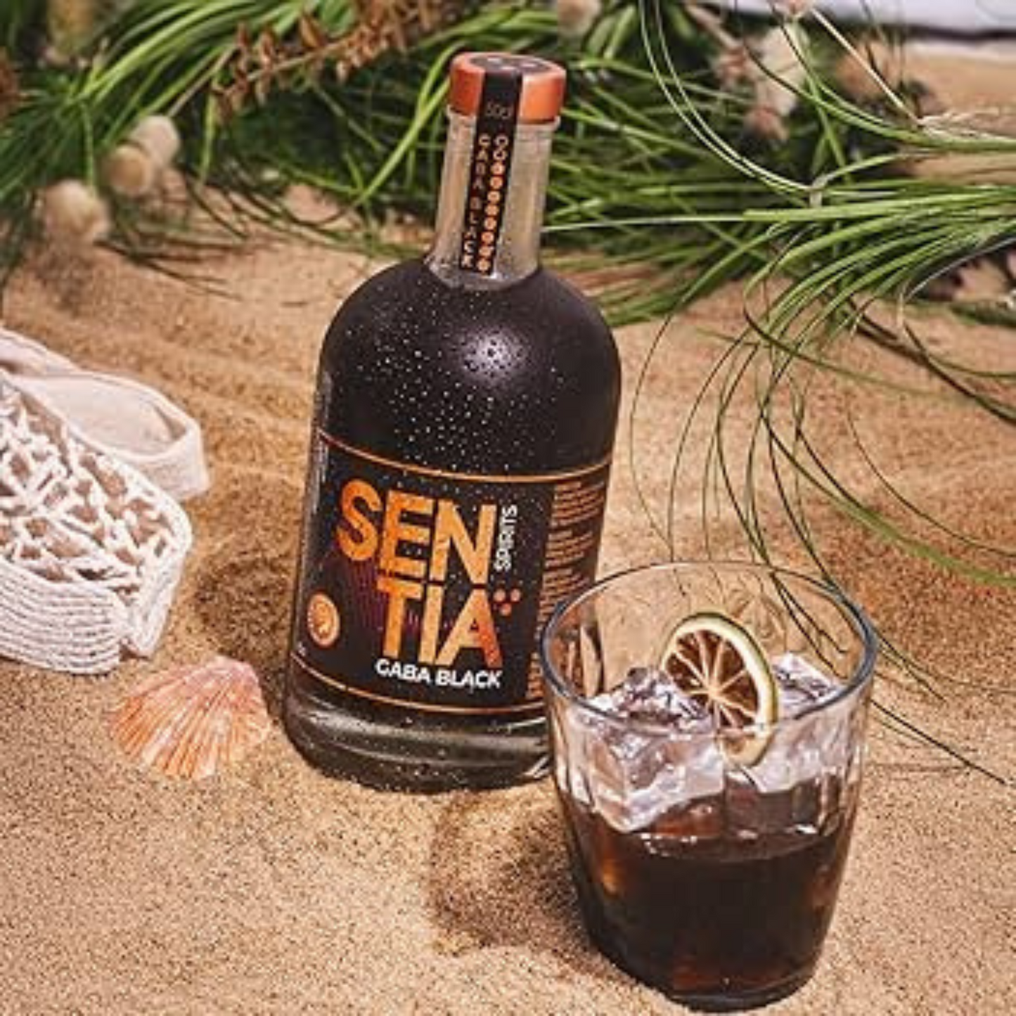 Bottle of Sentiia Caba Black rum on sand with a glass of dark liquid and ice.