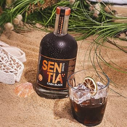 Bottle of Sentiia Caba Black rum on sand with a glass of dark liquid and ice.